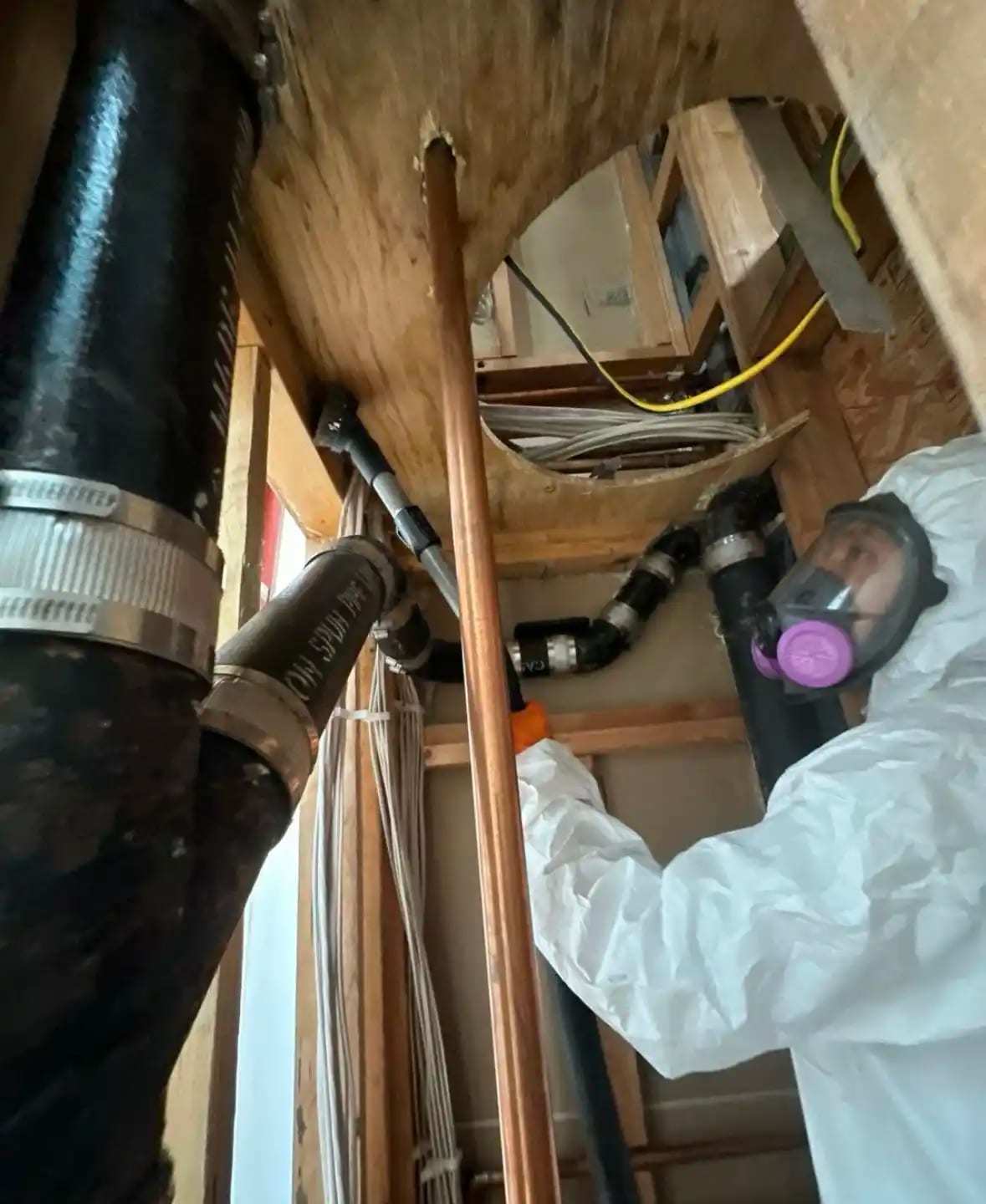 Technician in a respirator inspecting pipes inside an opened wall cavity